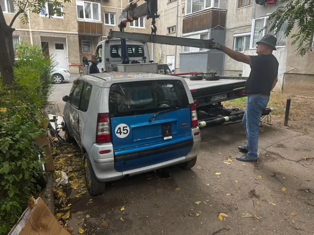 Un bărbat în cămașă neagră și blugi deschizi ridică o bară metalică de pe platforma unui camion de remorcare lângă un autovehicul mic albastru și gri.