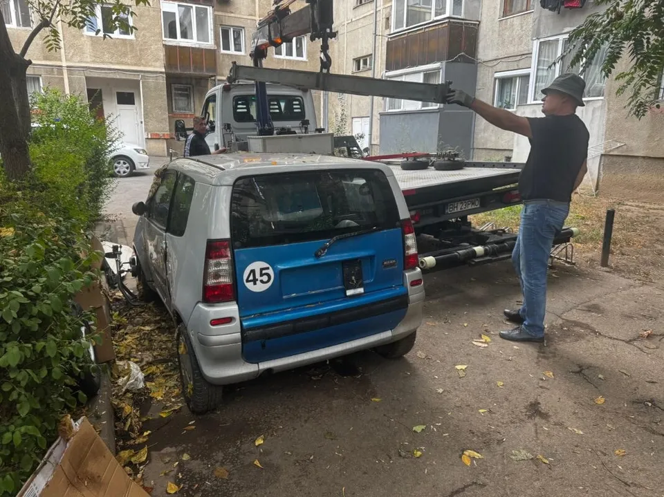 Un bărbat în cămașă neagră și blugi deschizi ridică o bară metalică de pe platforma unui camion de remorcare lângă un autovehicul mic albastru și gri.