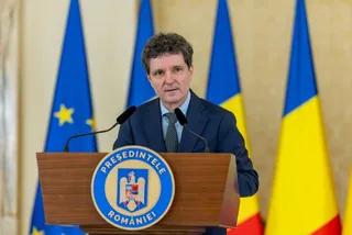 Un bărbat cu părul închis la culoare, purtând sacou și cravată, stă în spatele unui podium de lemn cu un emblema circulară în față.
