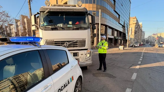 Un camion alb stă pe o stradă lângă o mașină de poliție locală, în timp ce un agent cu vestă reflectorizantă verde stă pe trotuar.