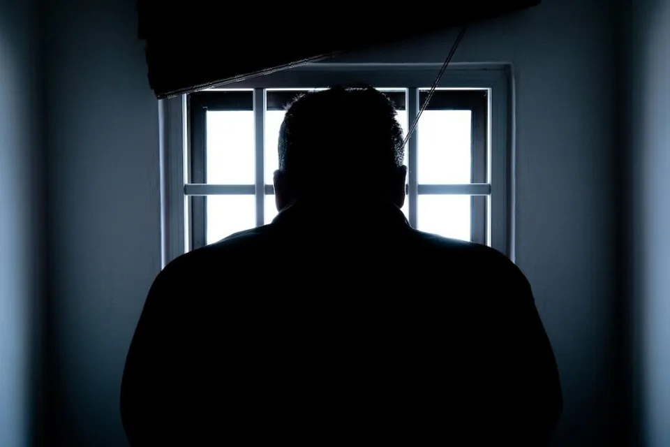 Silueta întunecată a unui bărbat stă cu spatele la cameră în fața unei ferestre prin care intră lumină albă.