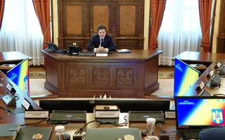 Un bărbat cu haine oficiale stă așezat la un birou de lemn în fața unor ferestre cu draperii portocalii și perdele albe.