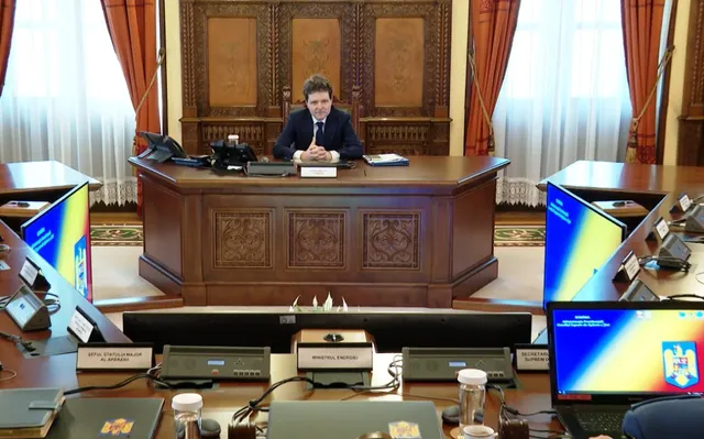 Un bărbat cu haine oficiale stă așezat la un birou de lemn în fața unor ferestre cu draperii portocalii și perdele albe.