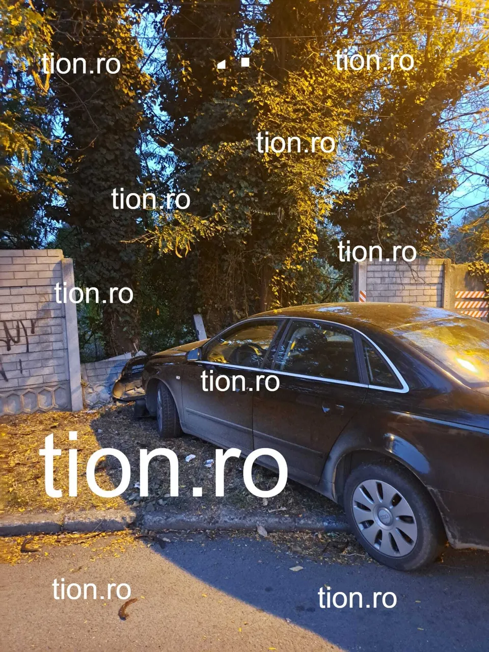 O mașină neagră este oprită pe marginea unui drum lângă un gard de beton și copaci sub lumina soarelui.