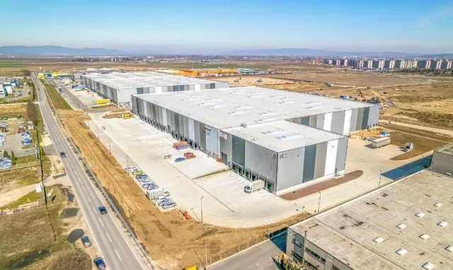 O vedere aeriană prezintă un ansamblu de clădiri industriale gri cu acoperișuri plate, situate lângă o șosea cu câteva vehicule și terenuri deschise.