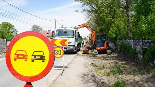 Un utilitar alb și o excavatoare portocalie execută lucrări pe un drum de lângă un gard și vegetație sub un cer senin.