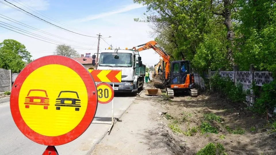 Un utilitar alb și o excavatoare portocalie execută lucrări pe un drum de lângă un gard și vegetație sub un cer senin.