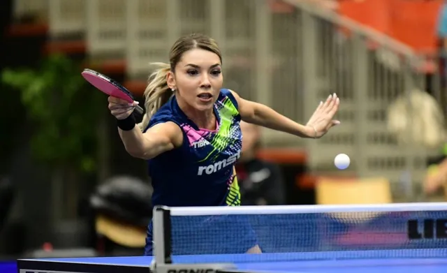 O femeie cu părul blond în echipament sportiv albastru lovește o minge de tenis de masă cu o paletă roz lângă o masă de joc.