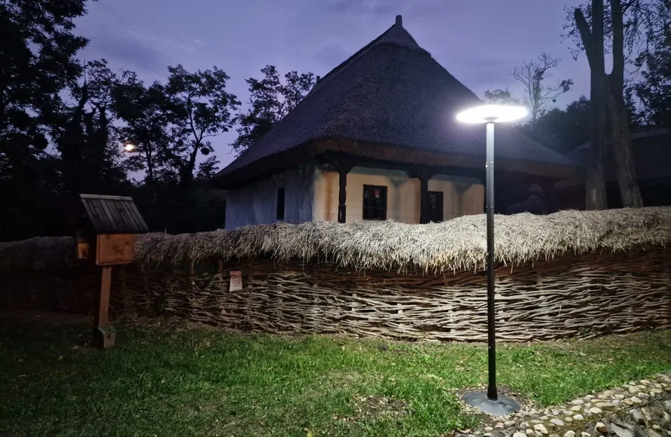 Un stâlp de iluminat modern este aprins în fața unei construcții tradiționale cu acoperiș de paie și un gard din răchită sub un cer nocturn.