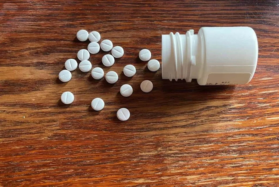 Diverse pastile albe rotunde sunt împrăștiate pe o suprafață de lemn lângă un flacon de plastic alb.