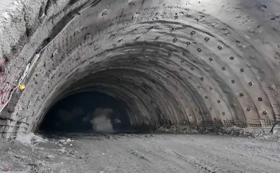 Interiorul unui tunel de beton cu pereți curbați și tavanul marcat de elemente de susținere metalice dispuse în rânduri regulate pe suprafața gri.