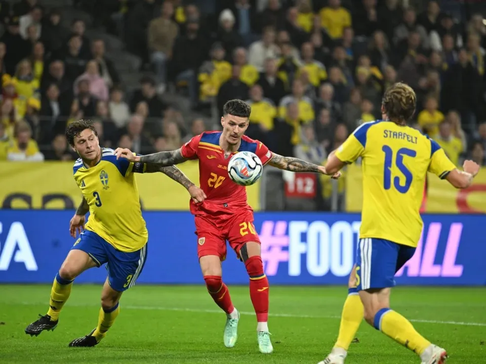 Trei jucători de fotbal sunt pe teren de joc, unul purtând echipament roșu și alții doi purtând echipament galben și albastru.