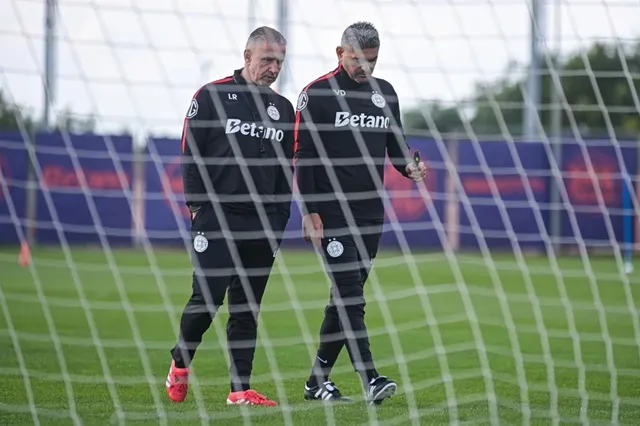 Doi bărbați îmbrăcați în echipamente sportive negre cu detalii albe și roșii merg pe un teren de fotbal prin spatele unei plase de antrenament.
