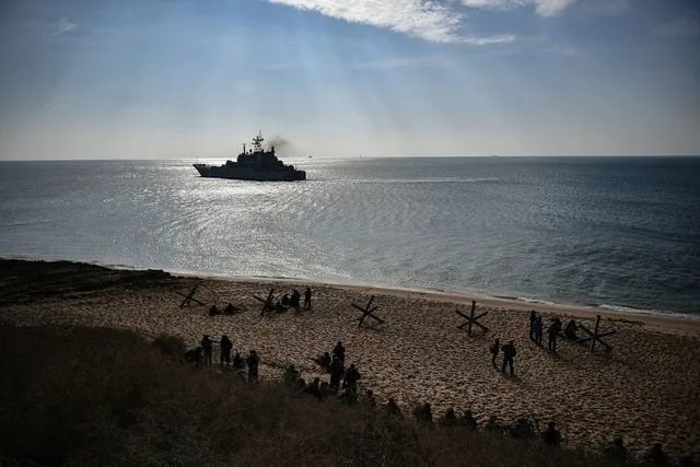 O navă militară navighează pe o mare calmă sub raze de lumină solare, în timp ce persoane stau pe o plajă nisipoasă în prim-plan.