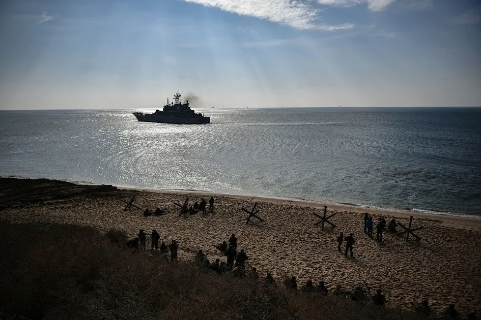 O navă militară navighează pe o mare calmă sub raze de lumină solare, în timp ce persoane stau pe o plajă nisipoasă în prim-plan.