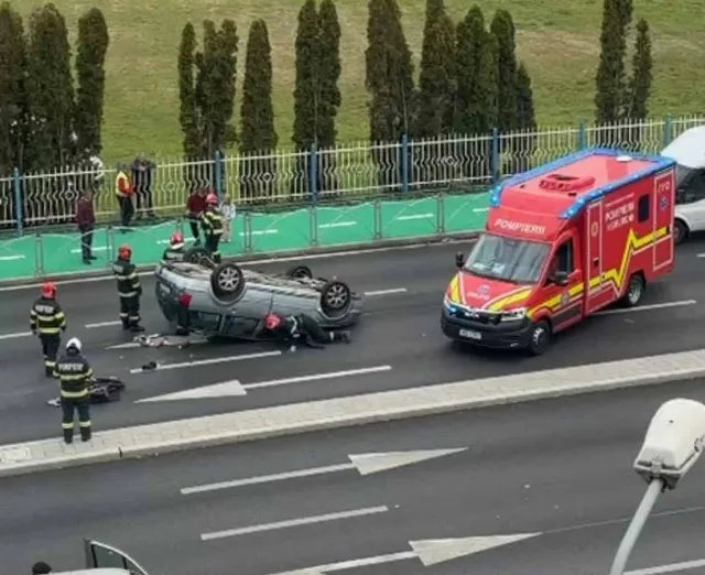 O mașină gri este răsturnată pe lateral pe un drum asfaltat, lângă o ambulanță roșie și galbenă și mai mulți salvatori în uniforme fluorescente.
