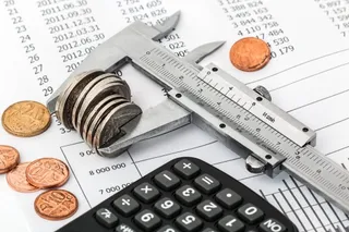 Un clește de măsurat metalic, câteva monede și un calculator negru sunt așezate pe un document cu tabele și cifre.