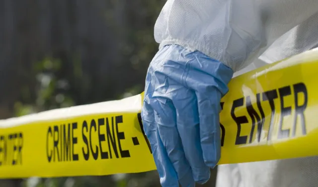 O persoană purtând mănușă albastră și costum alb ține o bandă galbenă pe care scrie text negru cu cuvintele crime scene și enter.
