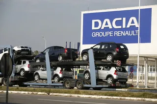 Mai multe mașini de culori diferite sunt transportate pe un remorcă de tip platformă pe un drum asfaltat.