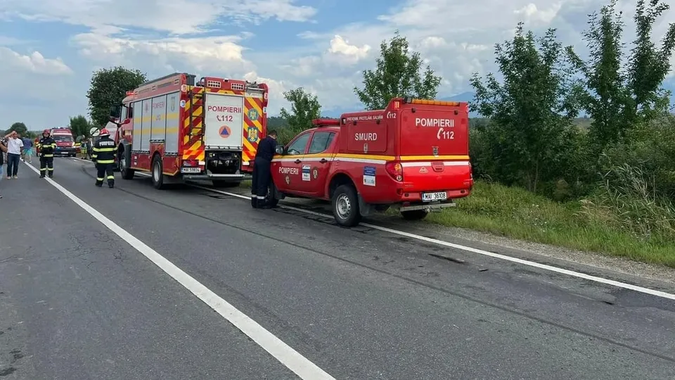 Vehicule de serviciu de culoare roșie, inclusiv un camion de pompieri și o mașină de intervenție, sunt oprite pe marginea unei drumuri asfaltate.