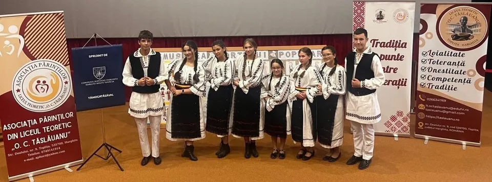 Un grup de opt tineri stă în rând pe o scenă purtând costume populare albe cu detalii negre și maro, lângă diverse panouri informative colorate.