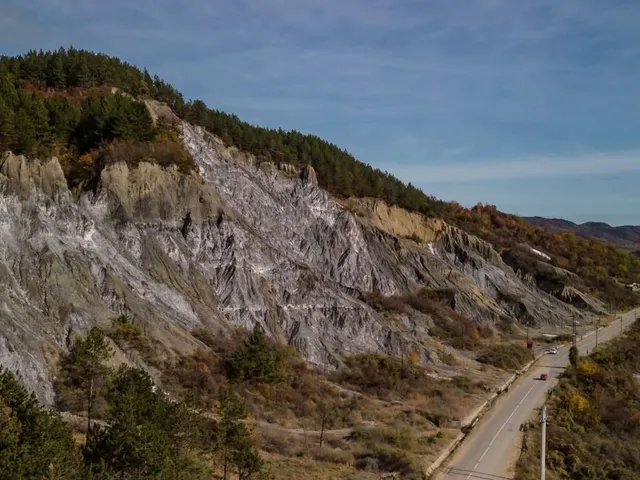 O imagine panoramică cu un drum pavat care serpentinează pe lângă o stână cu aspect de râpa și pădure pe deal.