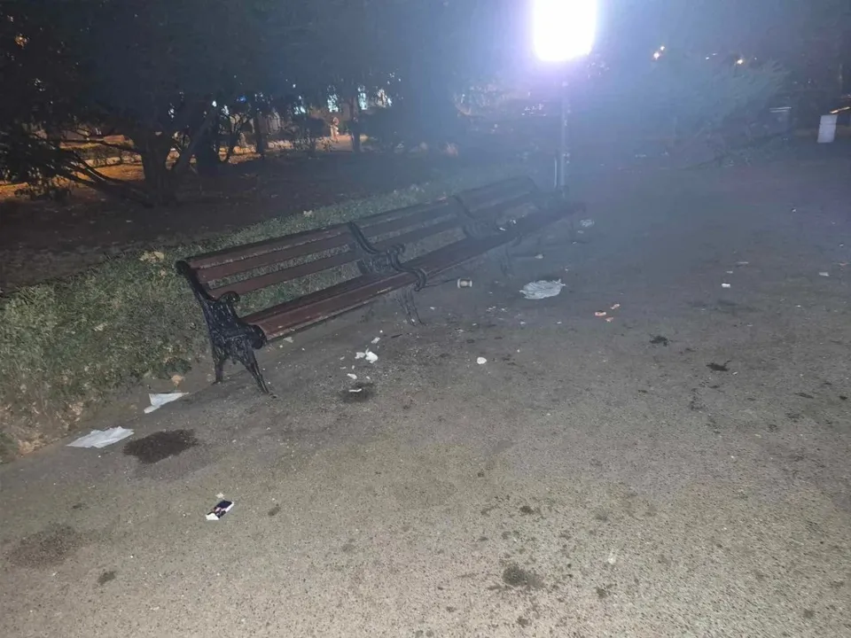 Un banc de parcul din lemn și metal este situat pe un drum de pietriș presărat cu diverse obiecte albe de deșeuri sub lumină nocturnă.