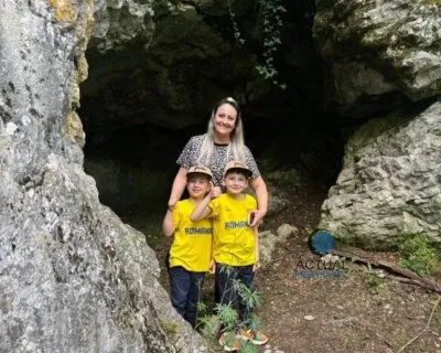 O femeie stă în spatele a doi copii care poartă tricouri galbene în fața unei peșteri mari de piatră.