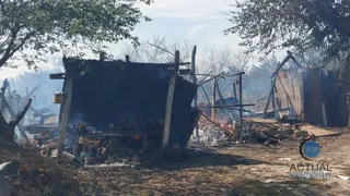 O structură de lemn carbonizată și o anexă parțial distrusă se află pe un teren cu resturi și cenușă sub arbori.