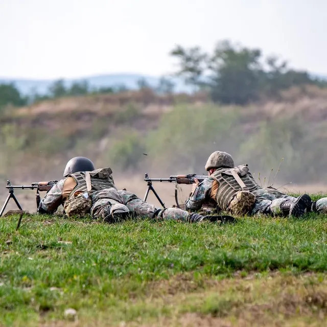 Două persoane purtând uniforme de camuflaj și căști sunt întinse pe iarbă, folosind arme cu bipod într-un teren deschis.