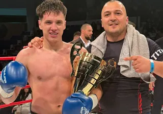 Un bărbat fără tricou cu mănuși de boxer albastre ține un trofeu auriu lângă un alt bărbat care poartă un prosop pe umeri.