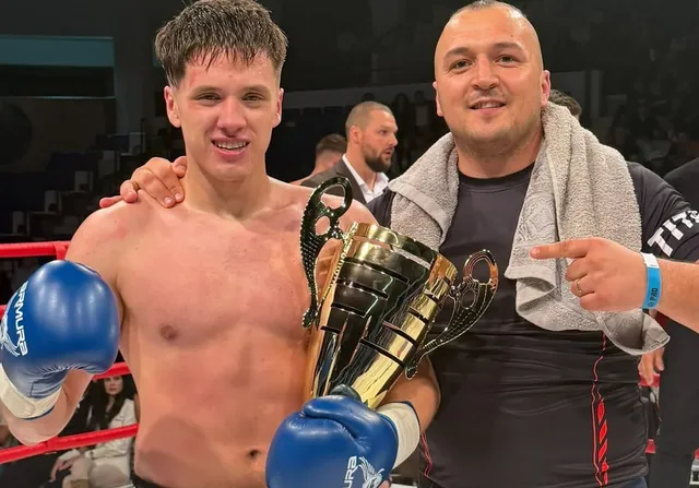 Un bărbat fără tricou cu mănuși de boxer albastre ține un trofeu auriu lângă un alt bărbat care poartă un prosop pe umeri.