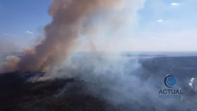 Un nor dens de fum maro și alb se ridică deasupra unui peisaj de vegetație întunecată sub un cer senin.