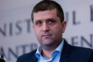 Un bărbat cu părul scurt și cămașă albastră sub un sacou închis la culoare privește spre lateral în fața unui fundal cu litere întunecate.