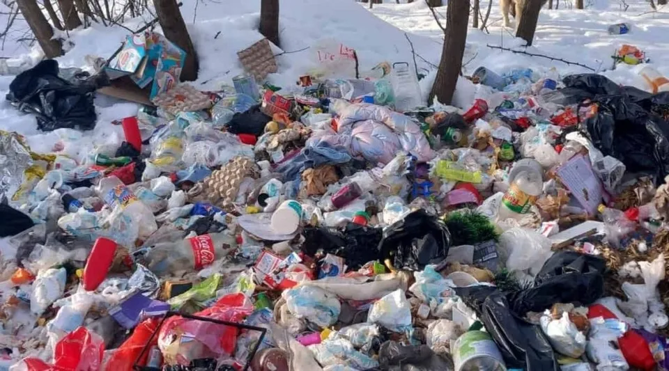 Un morman mare de deșeuri, inclusiv saci de plastic, ambalaje și resturi diverse, este împrăștiat pe un sol acoperit de zăpadă într-o zonă cu copaci.