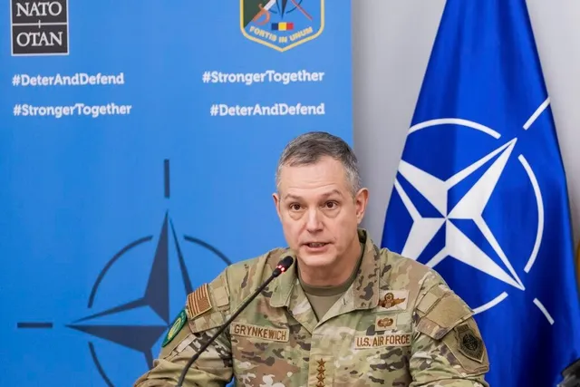 Un bărbat în uniformă militară de tip camuflaj stă în fața unui panou albastru cu logoul NATO și al unui steag albastru.
