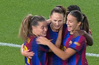 Patru femei purtând echipamente sportive albastre și roșii se îmbrățișează pe un teren de fotbal cu iarbă verde.