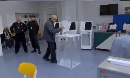 Un bărbat introduce un buletin într-o urnă transparentă, lângă cabine albe și doi bărbați în uniforme închise.