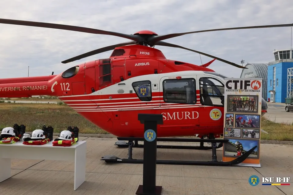 Un elicopter roșu și alb cu inscripția SMURD stă pe o pistă lângă un stand de prezentare și o masă cu echipamente de protecție.