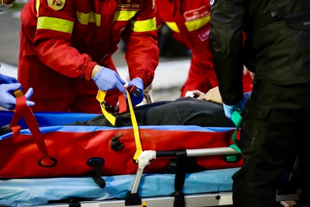 Persoane în uniforme roșii și negre transportă o persoană întinsă pe o targă roșie și albastră folosind chingi galbene.