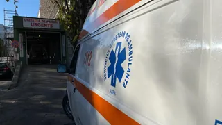 O ambulanță albă cu dungi portocalii și un simbol albastru este parcată în fața unei unități de primiri urgențe sub un cer senin.