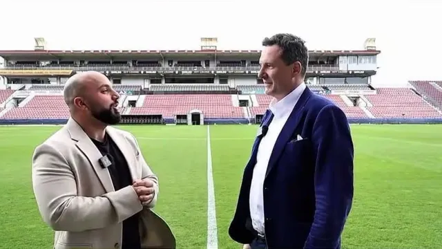 Doi bărbați stau pe un teren de fotbal verde, unul purtând un sacou bej și celălalt un sacou albastru peste o cămașă albă.
