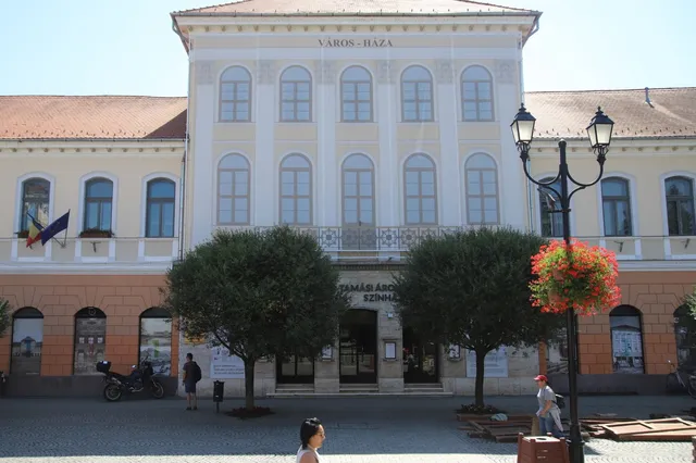 O clădire cu ferestre înalte și un balcon central este situată în spatele a doi copaci rotunzi și unui stâlp de iluminat cu flori.