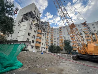 O macaraare galbenă stă lângă un bloc de apartamente cu o porțiune de fațadă și etaje prăbușite, lăsând să se vadă interiorul camerelor.