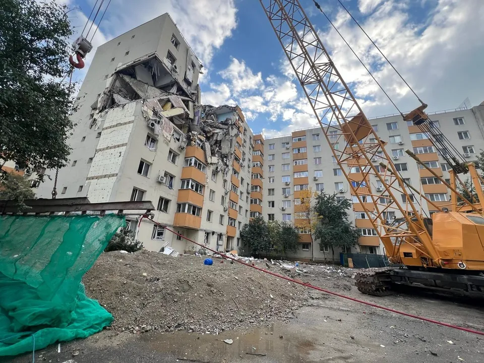 O macaraare galbenă stă lângă un bloc de apartamente cu o porțiune de fațadă și etaje prăbușite, lăsând să se vadă interiorul camerelor.