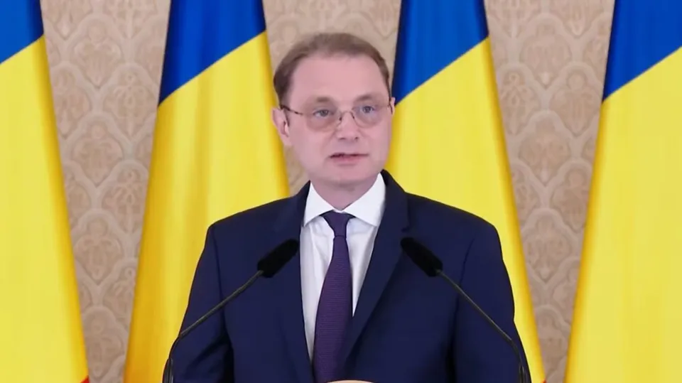 Un bărbat cu ochelari și cravată purtând un costum închis stă în fața unor steaguri galben și albastru.