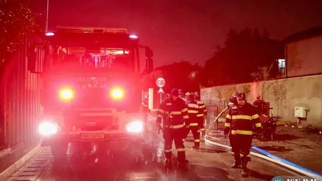 Patru pompieri în uniforme cu benzi reflectorizante merg pe un drum umed lângă un autospecială de pompieri cu luminile aprinse noaptea.
