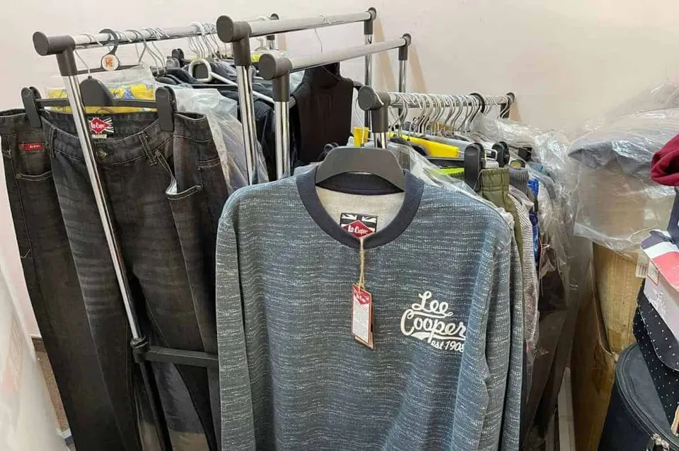 Haine și pantaloni de denim sunt suspendate pe umerașe pe două suporturi metalice de tip stender într-o încăpere.