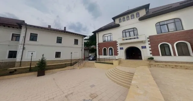 O clădire cu fațadă albă și secțiuni din cărămidă roșie este vizibilă lângă o curte pavată cu trepte largi și un gard metalic scurt.