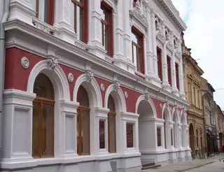 O clădire cu fațadă albă și roșie prezintă ferestre înalte, arcade și elemente decorative sculptate pe parcursul nivelurilor superioare și inferioare.